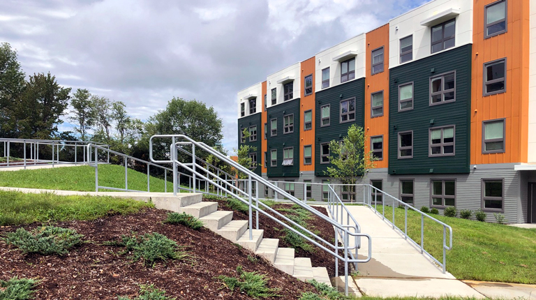 A white, green, and orange student housing building is bordered by accessible paths and green space. 