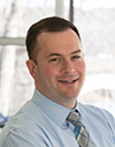 A man in a light blue shirt and blue, gray, white striped tie. 