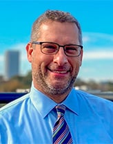 A business headshot of a man with brown hair wearing glasses, blue shirt and tie.