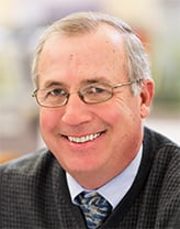 Joe Herr smiling, wearing a shirt, tie, and vest. 