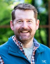 Headshot of Ryan Scott wearing a button-down business shirt and vest and smiling in an outdoor setting.