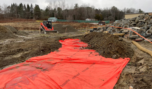 Orange plastic sheet partially covered with dirt in excavated area with bulldozer and pile of tires in background.