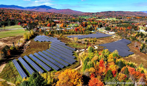 Aerial view of three large clusters of solar panels surrounded by fall foliage with mountains in the distance.