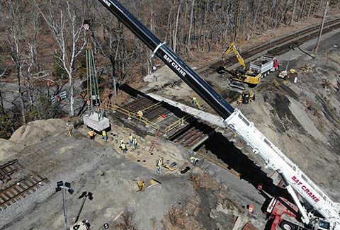 Workers use a crane and other heavy equipment to perform construction on an MBTA rail bridge over a street next to a body of water.