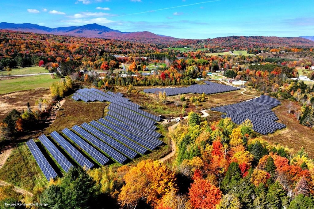 Aerial view of three large clusters of solar panels surrounded by fall foliage with mountains in the distance.