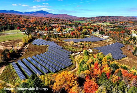 Aerial view of three large clusters of solar panels surrounded by fall foliage with mountains in the distance.