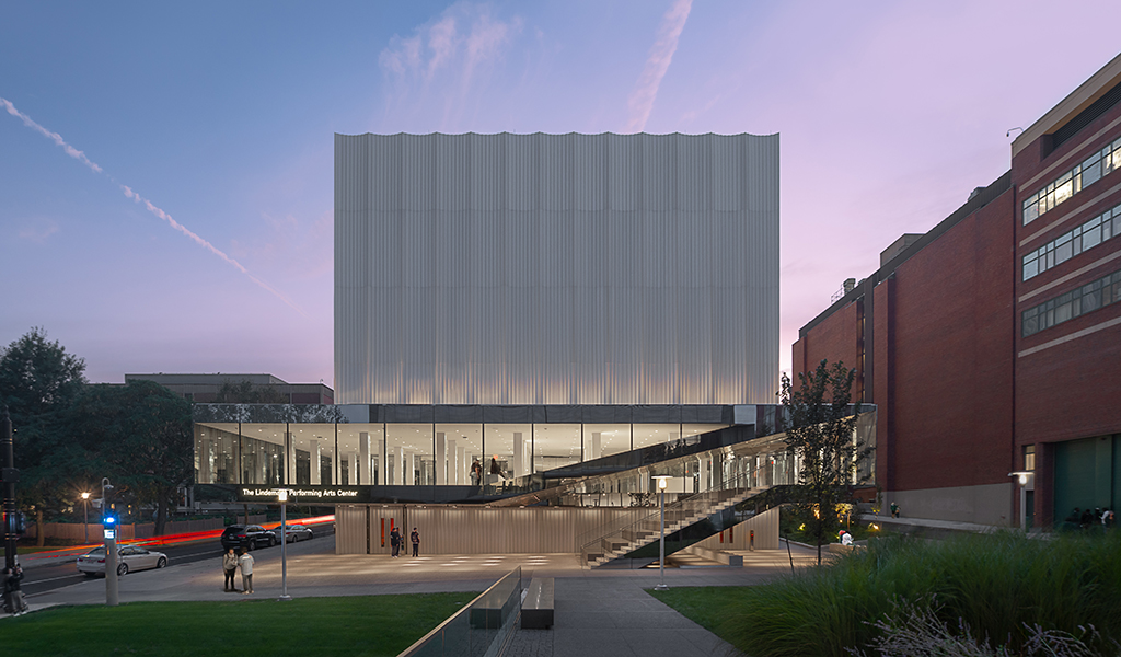 A digital rendering of Brown University’s new Performing Arts Center surrounding a green space with trees and students walking across a crosswalk.