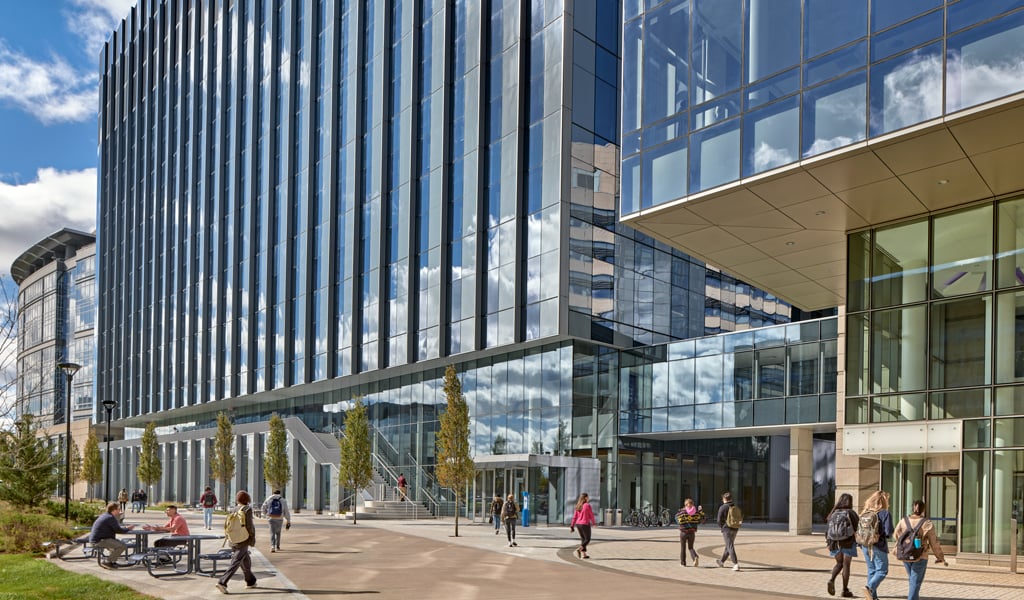 People walking and in conversation in the courtyard of two modern glass buildings with white staircase and connecting sky bridge.