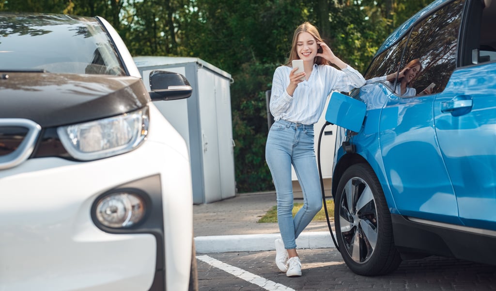 Young woman leaning on electric vehicle while it charges.