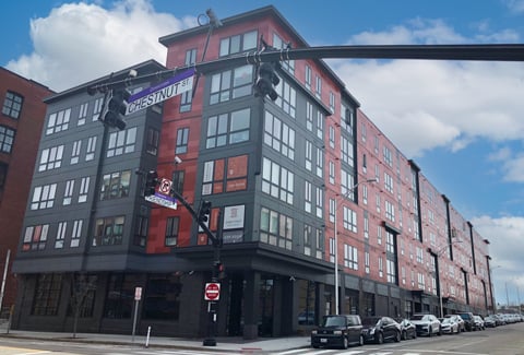 Urban street view of a modern six-story building featuring red and gray exterior and parked cars along sidewalk.
