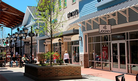 A small tree in a brick planter is in the middle of a walkway between rows of shops at Merrimack Premium Outlets.