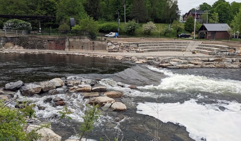 Numerous kayakers in multi-colored kayaks paddle in river rapids near a spectator viewing area.