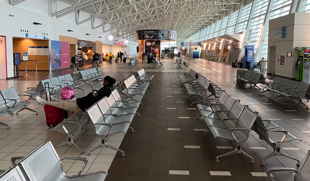 Passenger airport terminal with arched glass and steel ceiling, rows of seating, information signs, and travelers.