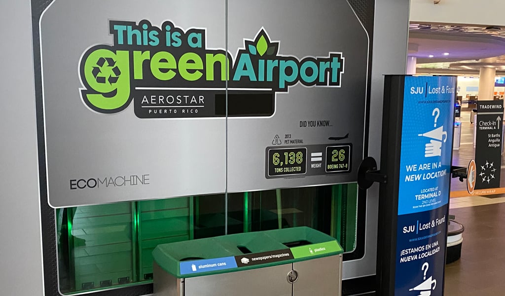 Gray, green and black signage depicting an airport’s commitment to sustainability hanging over recycling bins.