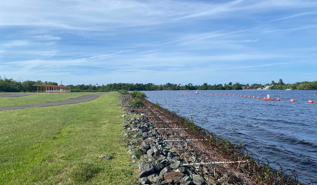 Body of water with orange buoys bordered by a rocky embankment, grassy area and paved path on the left, under a blue sky.