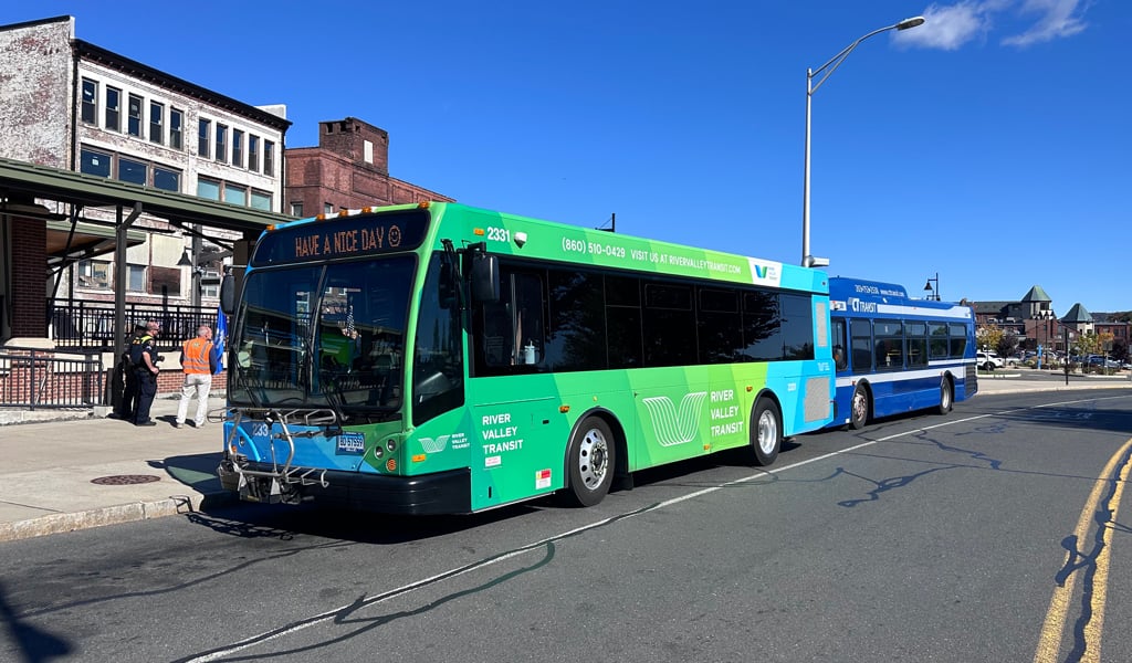 Green and blue River Valley Transit bus parked on side of the road with blue skies and brick buildings behind it.