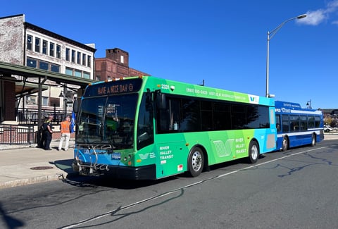 Green and blue River Valley Transit bus parked on side of the road with blue skies and brick buildings behind it.