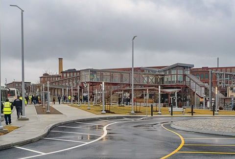 People gather and walk around the exterior of brick train station with glass walkways and windows.