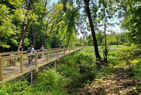 A proposed boardwalk running over a river showing bicyclists and pedestrians using the boardwalk through a wooded area.