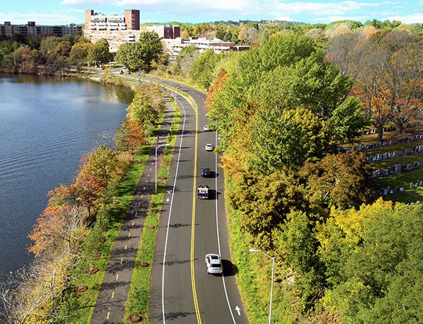 A two-lane roadway and bicycle path separated by green grass with a river to the left and trees and foliage to the right.