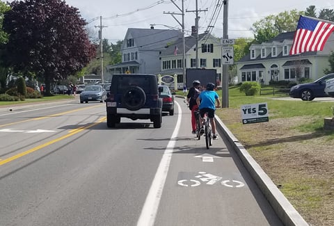 View of bike lane on Route 1 in Ogonquit, Maine, with two cyclists in the bike lane and multiple cars driving to their left.