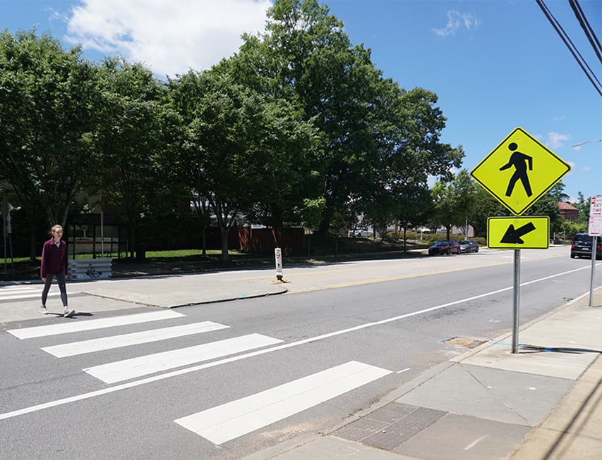 Pedestrian crossing in crosswalk with yellow pedestrian crosswalk sign on a roadway lined with green trees.