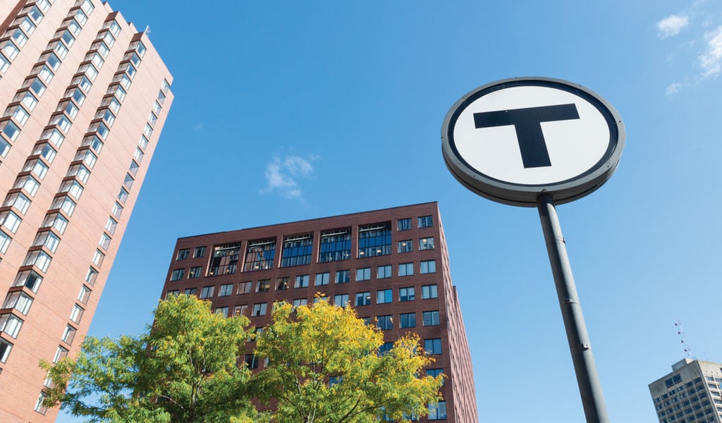 Cityscape with a prominent Boston MBTA transit system 'T' sign, surrounded by modern buildings and a clear blue sky.