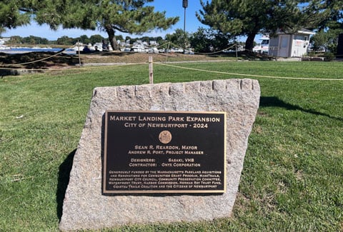 Commemorative plaque for the Market Landing Park Expansion displayed on a stone in a grassy area with trees and a blue sky.