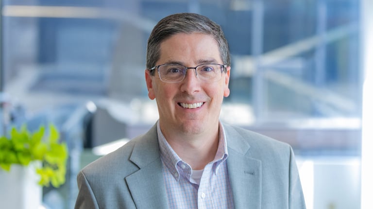 Chris Fronheiser wears glasses and a grey blazer in an office setting.