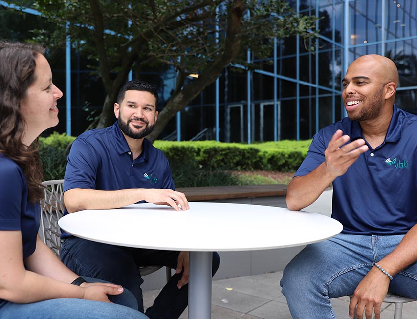 Three VHB team members sit talking around an outdoor table. 