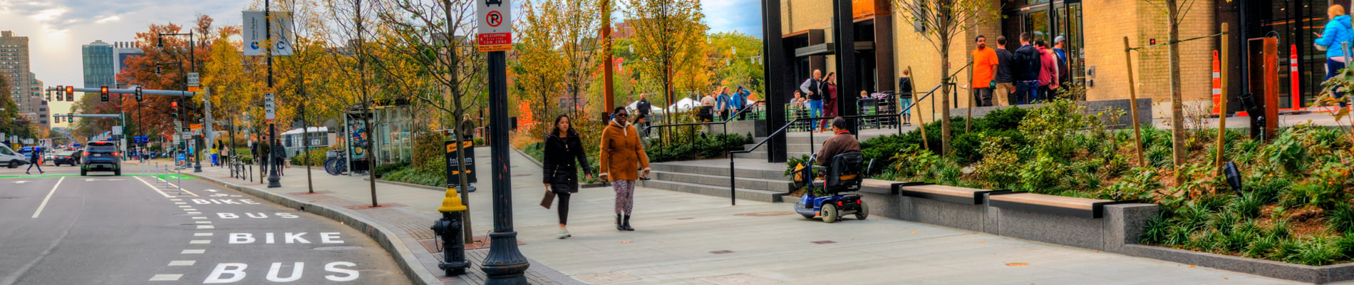 City streetscape with bike lanes, bus lanes, wide sidewalks, seat walls, and pedestrians.