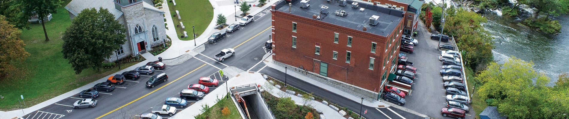 Aerial view of a small Vermont town with a historic church, brick buildings, a river waterfall, parked cars, and lush greenery. 