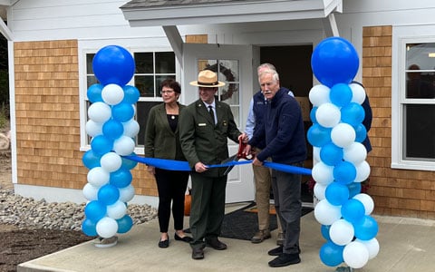 Three people participating in a ribbon-cutting ceremony in front of a new building, decorated with blue and white balloons. A camera is positioned in the foreground, capturing the event.