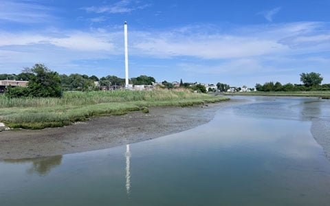 A scenic view of a coastal landscape featuring a tall white tower reflected in a tranquil water body, surrounded by lush greenery under a clear blue sky.