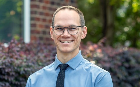 Bryce Corlett smiles at the camera wearing a blue shirt and glasses.