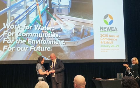 Two individuals shaking hands on stage at the NEWWA 2025 Annual Conference & Exhibit, with a backdrop displaying the slogan 'Work for Water. For Communities. For the Environment. For Our Future.' and images of water treatment infrastructure.