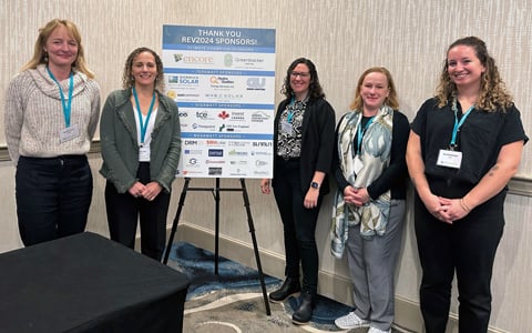 VHBer stand together wearing business attire in front of a sign thanking the 2024 REV Conference sponsors. 