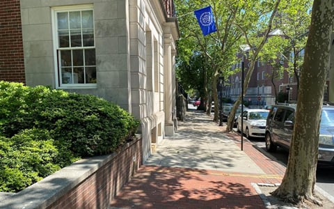 A sunny street view featuring a brick-lined sidewalk with trees providing shade, parked cars, and a building with a RISD flag hanging from a flagpole.