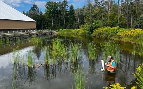 A person in a safety vest conducts water quality testing in a pond surrounded by lush greenery and wetland plants under a partly cloudy sky. A modern building is visible in the background.