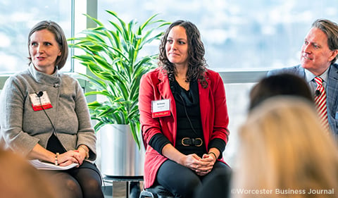 Three professionals attending a conference, seated with name tags, in a room with large windows and a cityscape in the background.