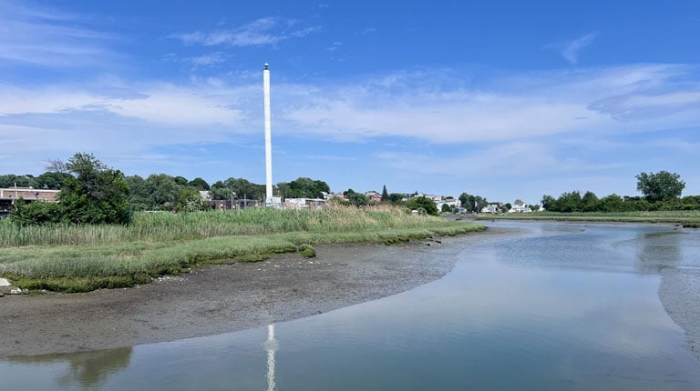 A scenic view of a coastal landscape featuring a tall white tower reflected in a tranquil water body, surrounded by lush greenery under a clear blue sky.