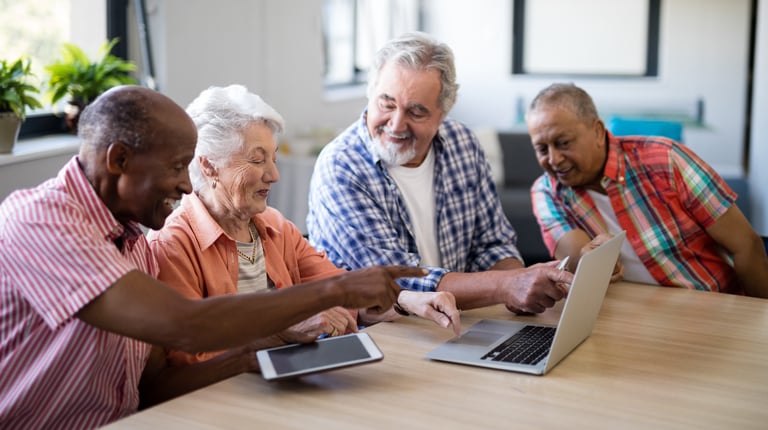 A group of elderly people are sitting at a table looking at a laptop computer and tablet together.