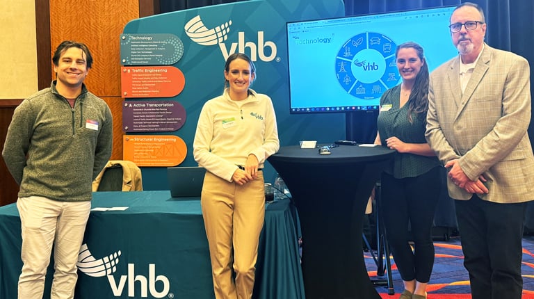 Four VHB employees standing in front of an exhibit booth featuring the VHB logo.