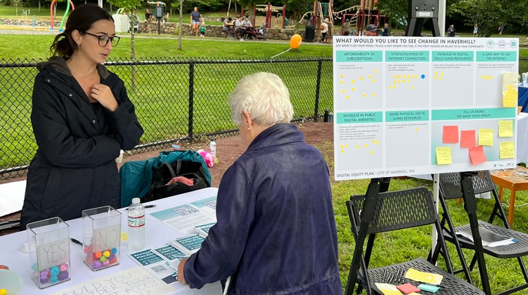 A VHB popup tent and table at the Farmers Market with a woman in a black coat speaking to a gentleman in a gray sweatshirt. 