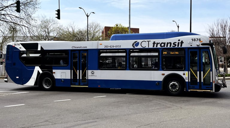 A bus stop sign with a CTtransit bus in the background on the other side of the street.