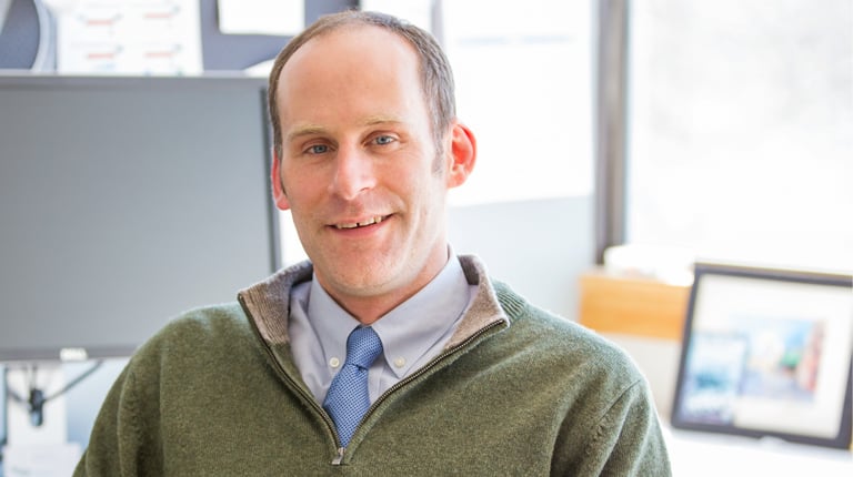 Peter Smiar seated in an office, wearing a sweater over a collared shirt. The background includes a window and a computer monitor with various notes attached.