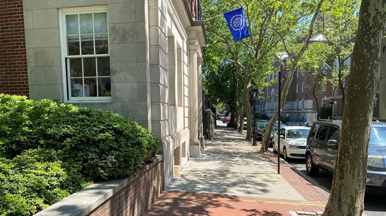 A sunny street view featuring a brick-lined sidewalk with trees providing shade, parked cars, and a building with a RISD flag hanging from a flagpole.