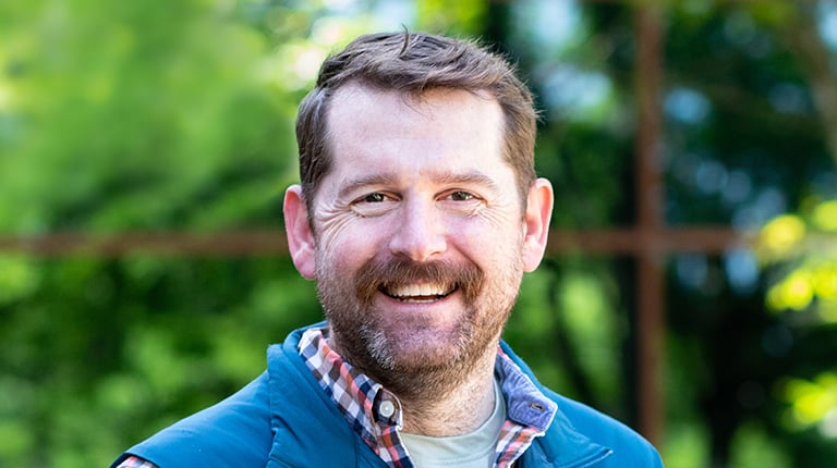 Headshot of Ryan Scott, a man wearing a plaid shirt and a vest smiling with trees in the background. 