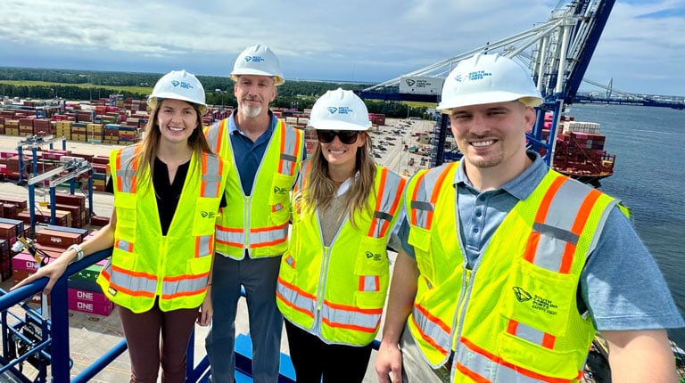 People wearing hard hats and standing on a crane above a port.