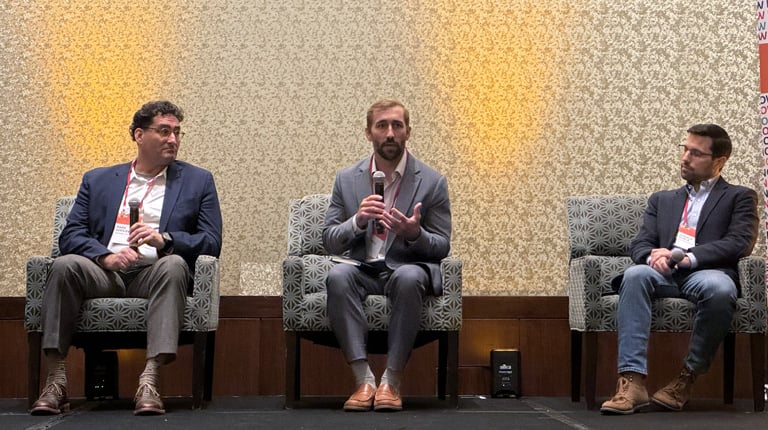 Three panelists, sitting on stage in a conference room, engage with the audience. The central panelist holds a microphone.